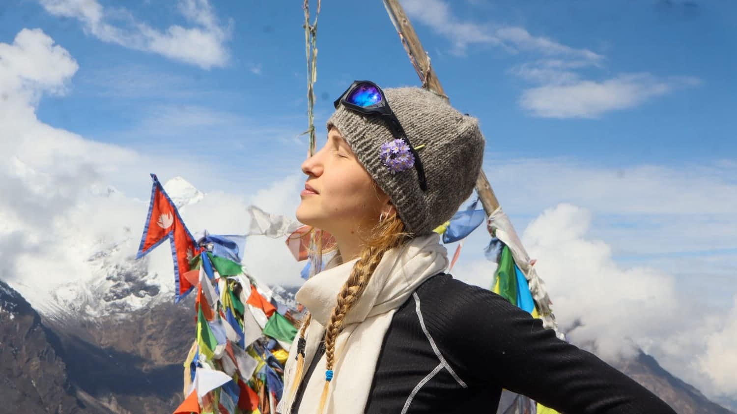 A woman in profile with her eyes closed faces the sun, the Nepali flag and prayer flags fluttering beside her.